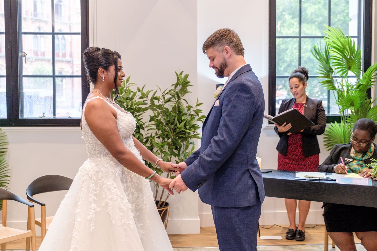 A couple holds hands during their wedding ceremony in a bright indoor venue with natural light and greenery.