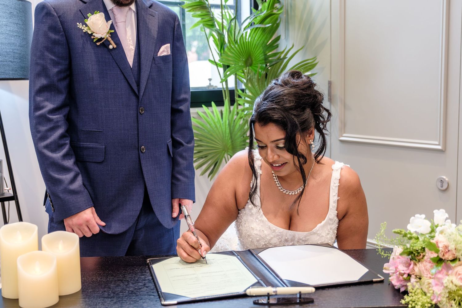 A couple signs wedding documents at a ceremony table decorated with blush flowers and white candles.