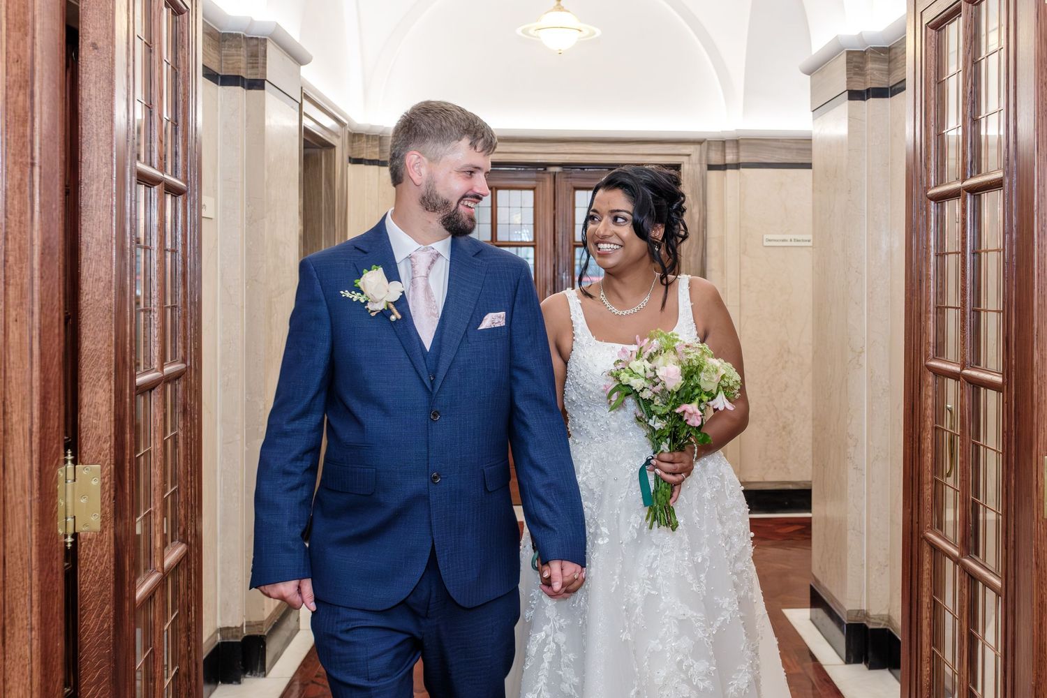 Elegant couple shares romantic moments in a grand hallway with wooden French doors and classic decor.