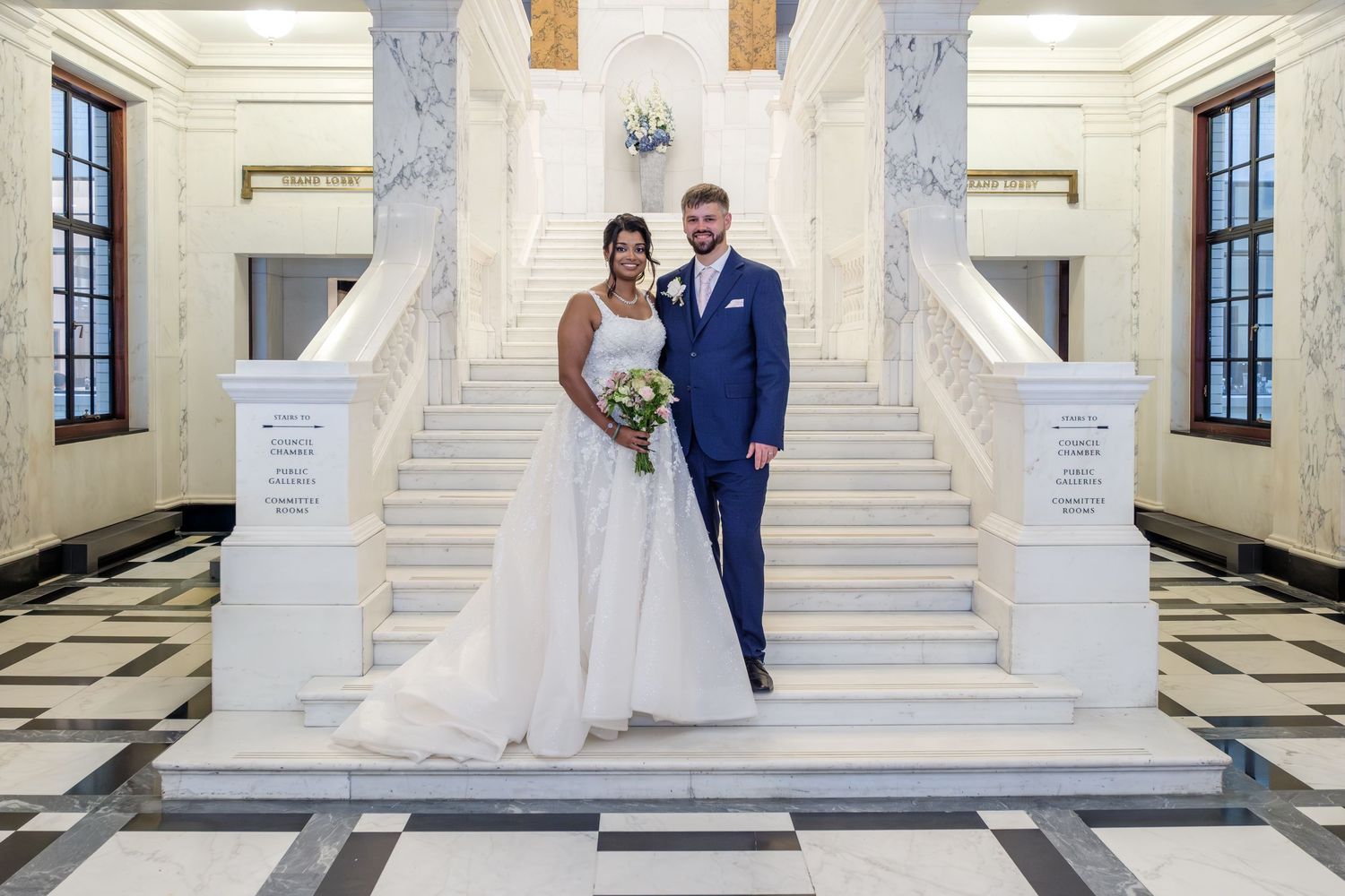 Elegant wedding couple poses on grand white marble staircase in traditional formal attire during their ceremony.