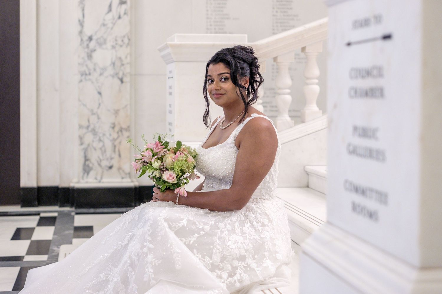 A bride in a white lace gown poses elegantly amidst white columns during a photo session.