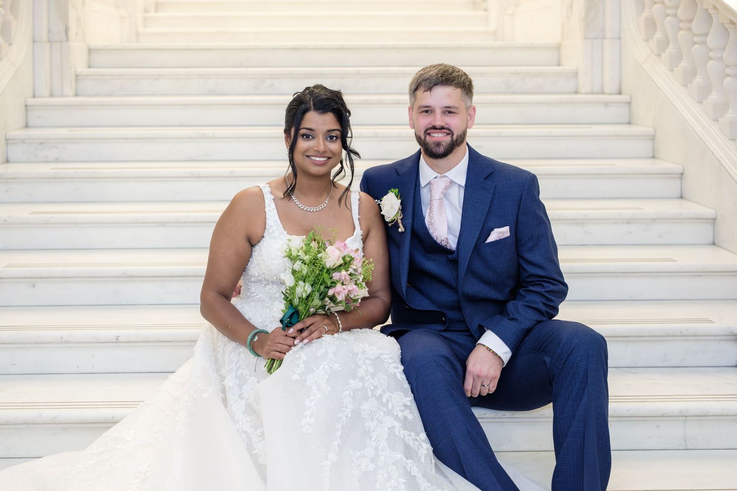 A cheerful wedding couple sits on white marble steps, the bride in a white gown holding a bouquet of yellow flowers.