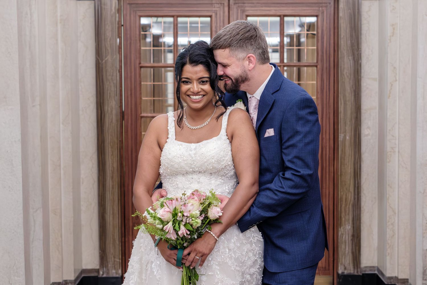 A happy newlywed couple sharing a romantic moment in front of wooden doors while holding a white rose bouquet.