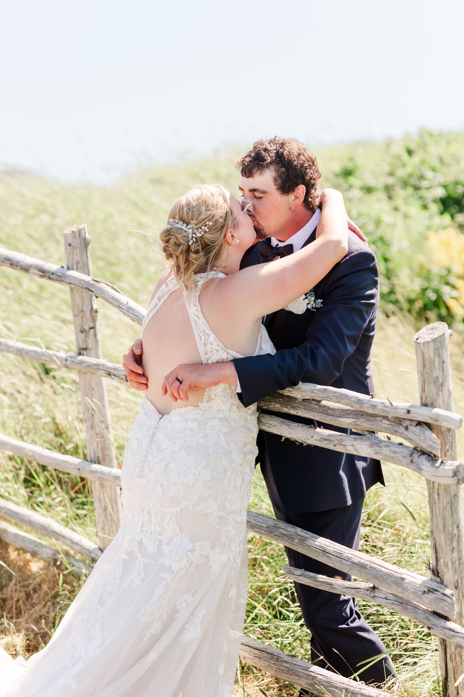 A romantic wedding couple embraces beside a rustic wooden fence in a sunny meadow.