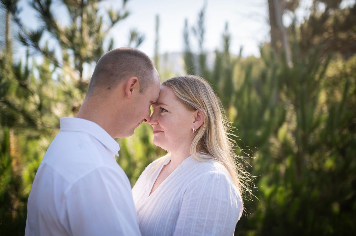A romantic couple in white outfits share an intimate moment together in front of greenery during a sunny outdoor photoshoot.