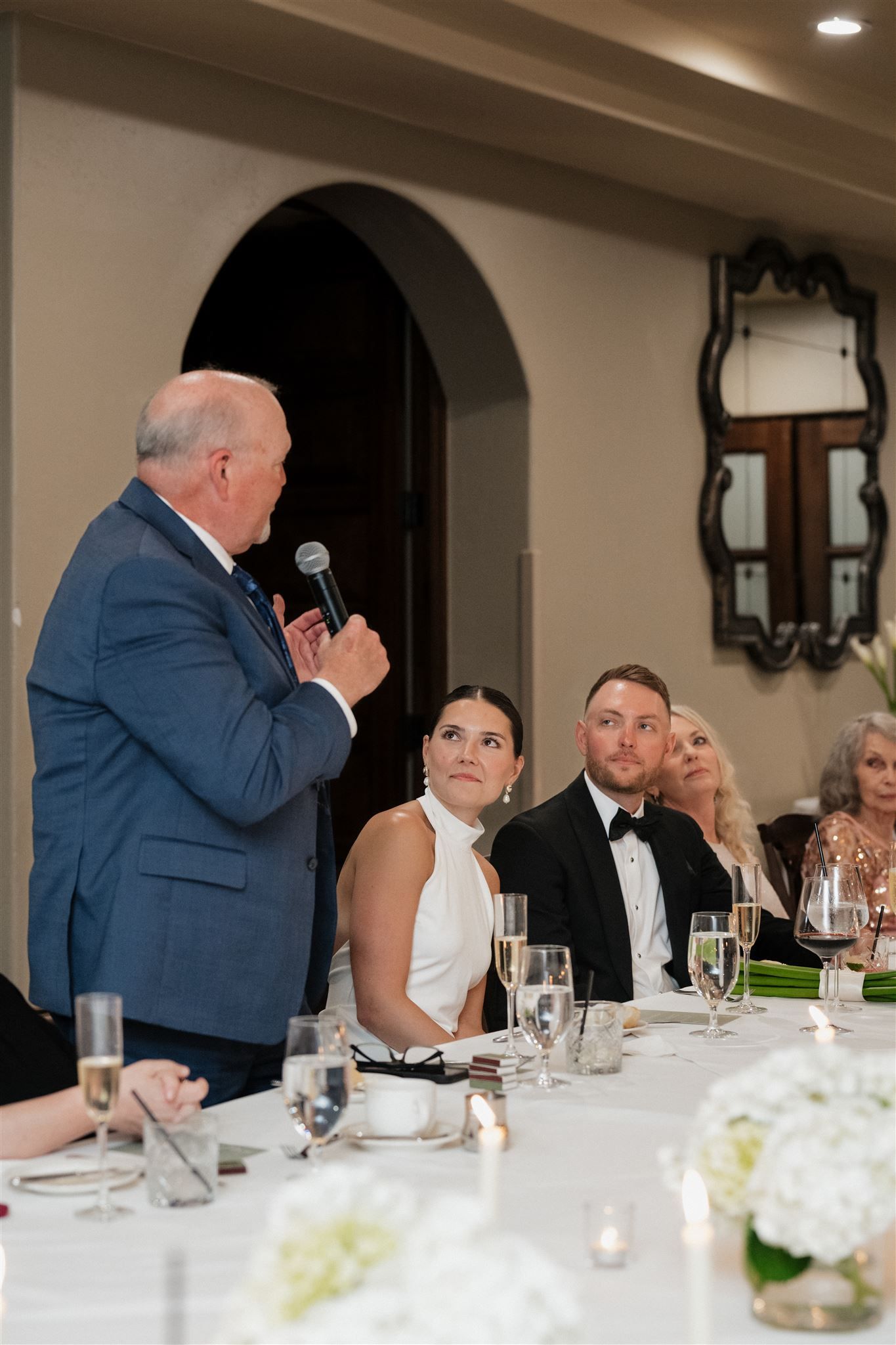 A speaker in a navy suit addresses guests during a formal dinner reception at a white-tablecloth event.