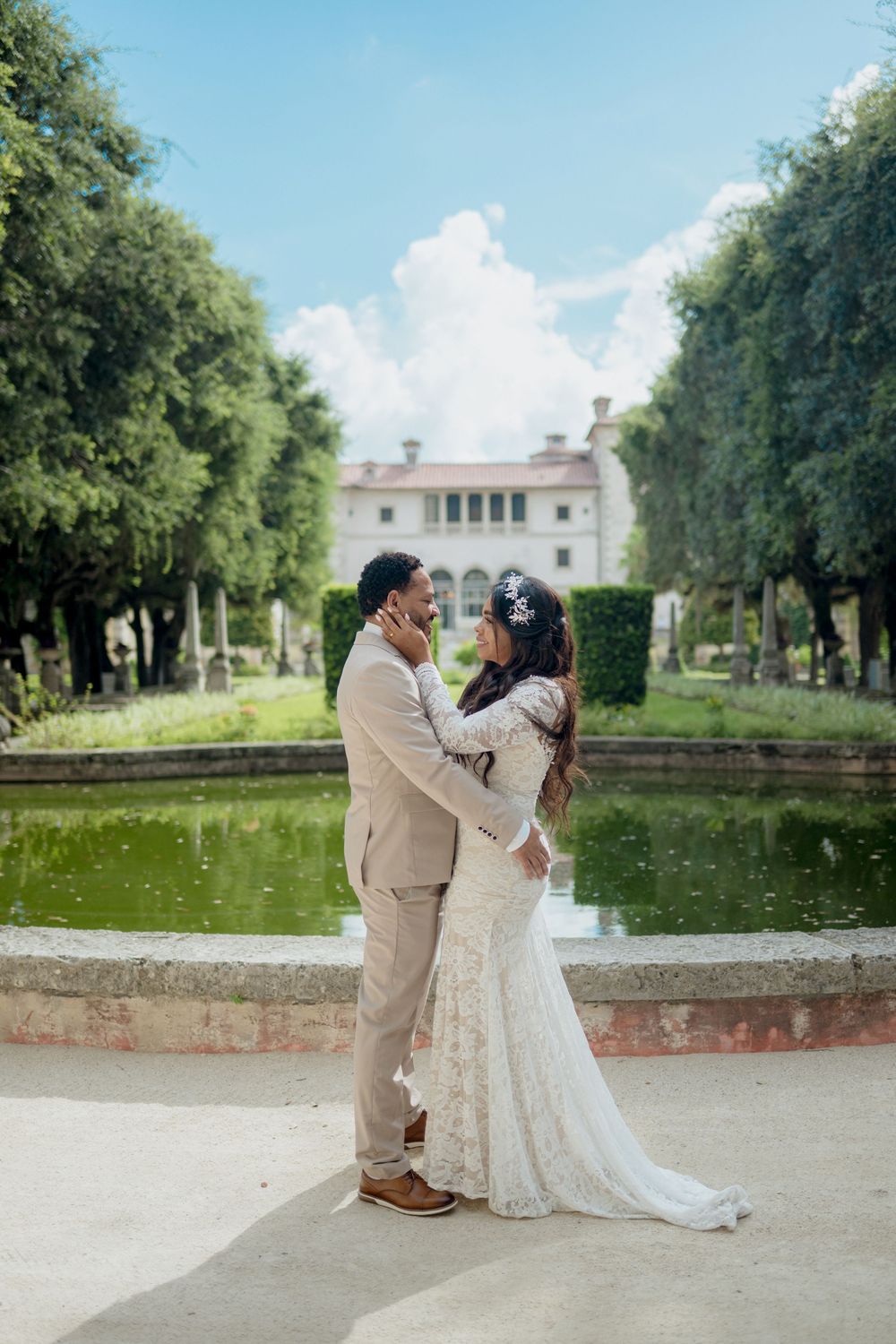 A romantic wedding photo of a couple embracing by a reflecting pool with a stately mansion and green trees in the background.