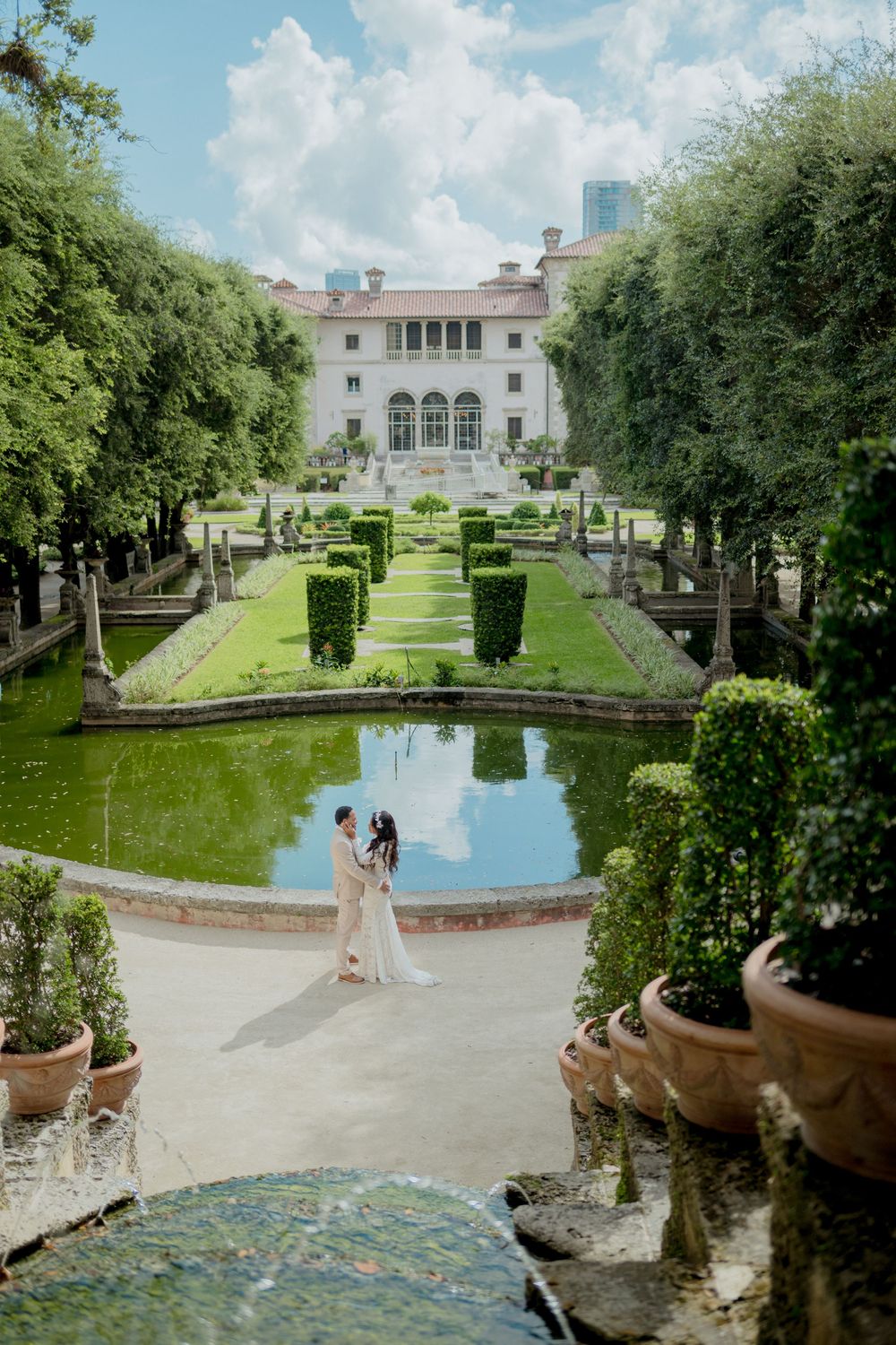 Elegant formal garden features a reflecting pool surrounded by manicured hedges and pathways leading to a grand mansion.