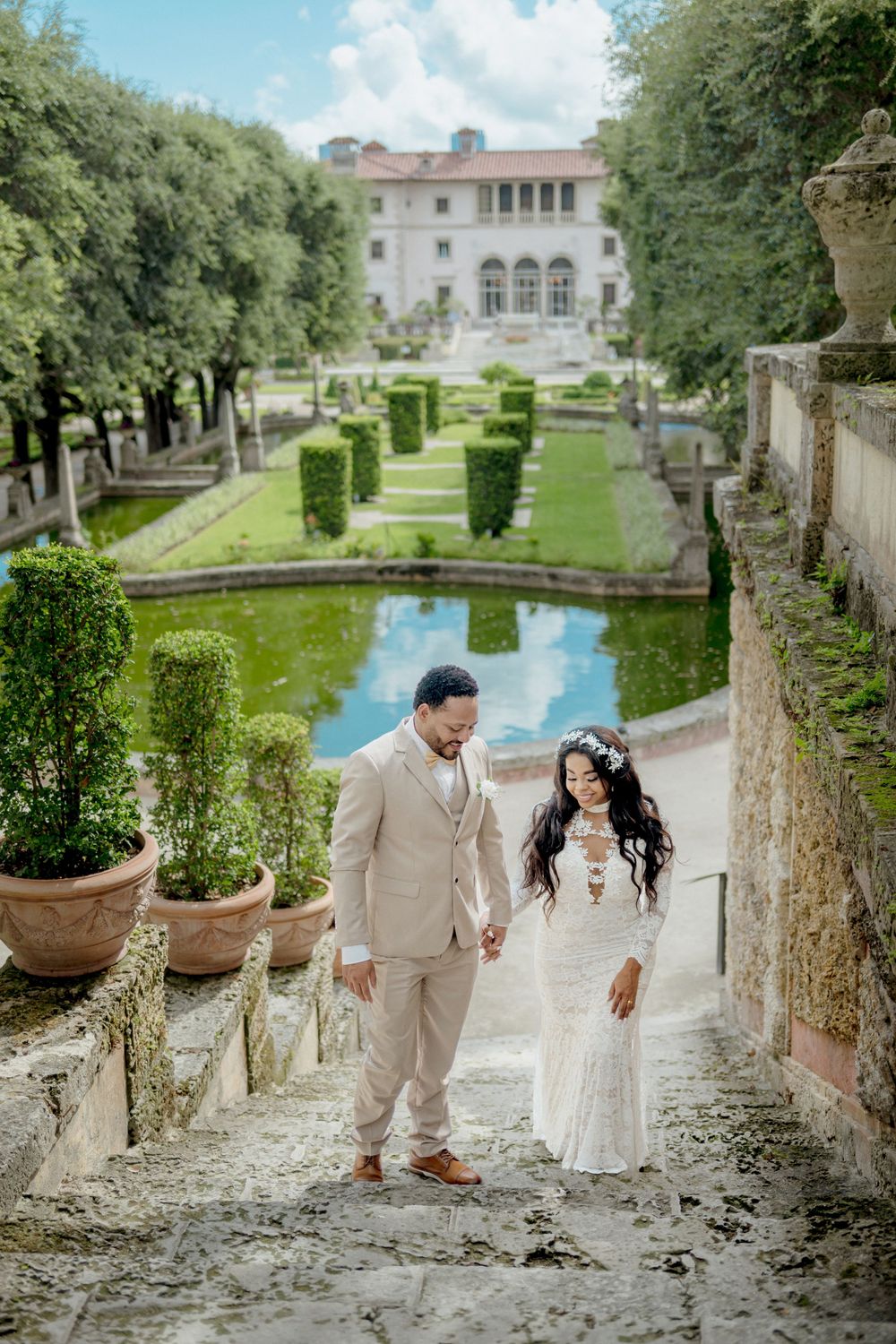 A couple stands on stone steps in front of a manicured garden with reflecting pool at an elegant Italian villa.