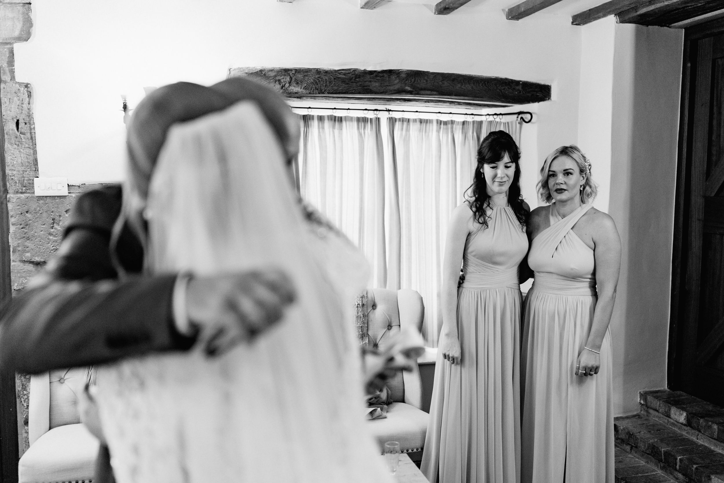 Two bridesmaids in matching gray dresses watch an emotional wedding moment in a rustic venue.