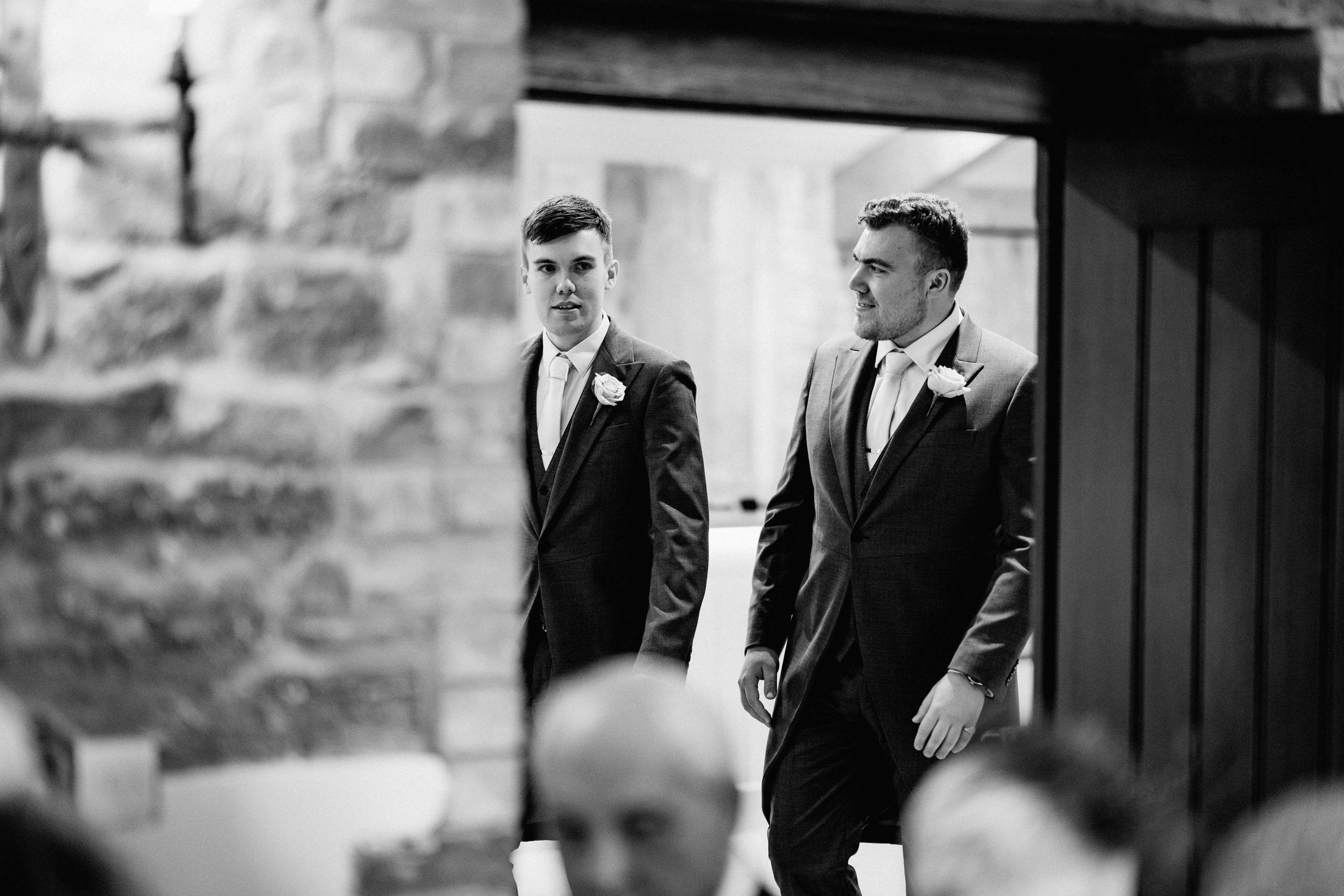 Two groomsmen in dark suits stand together during a formal wedding ceremony.