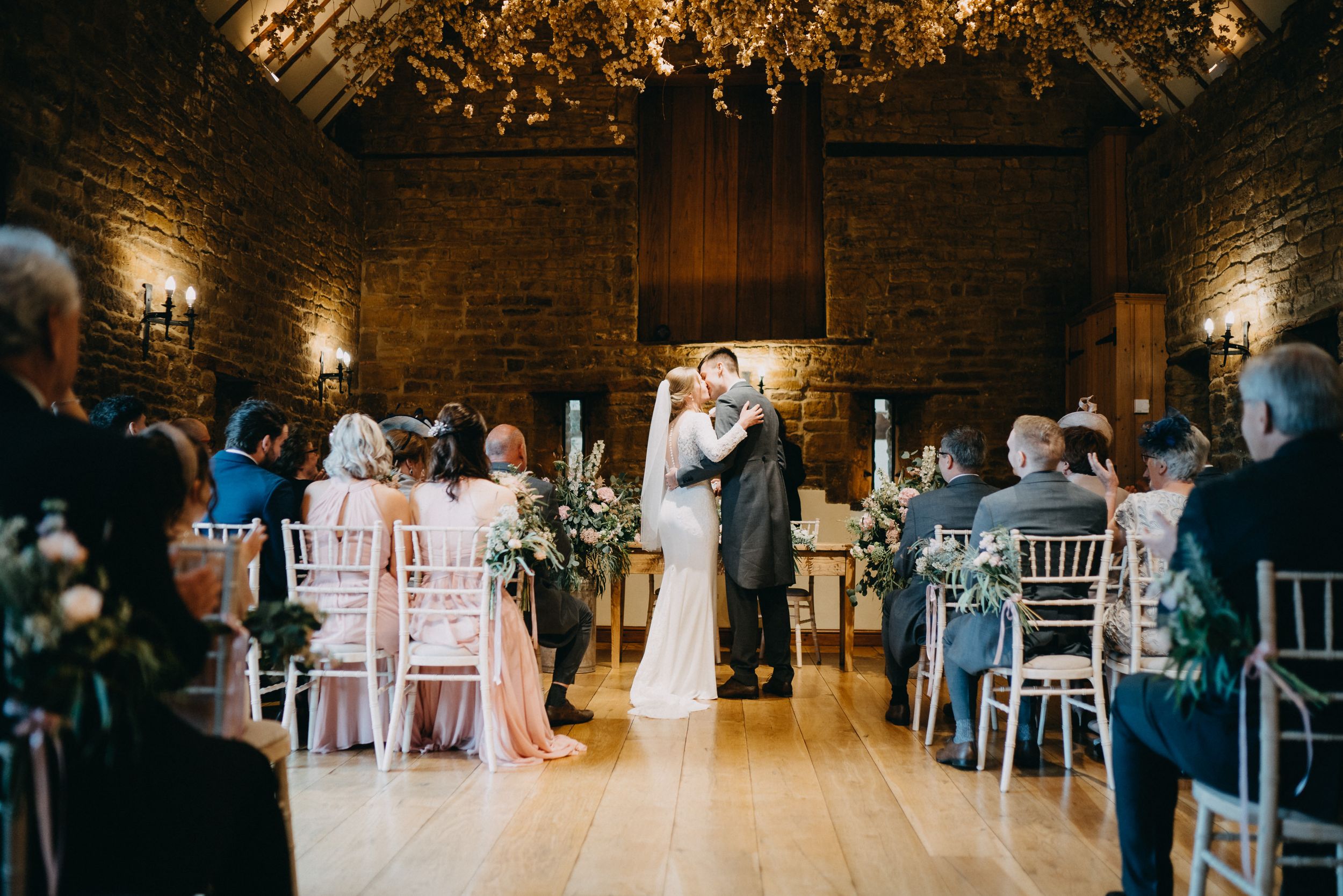 First kiss moment at Crockwell Farm in rustic brick venue with guests watching from white chairs.