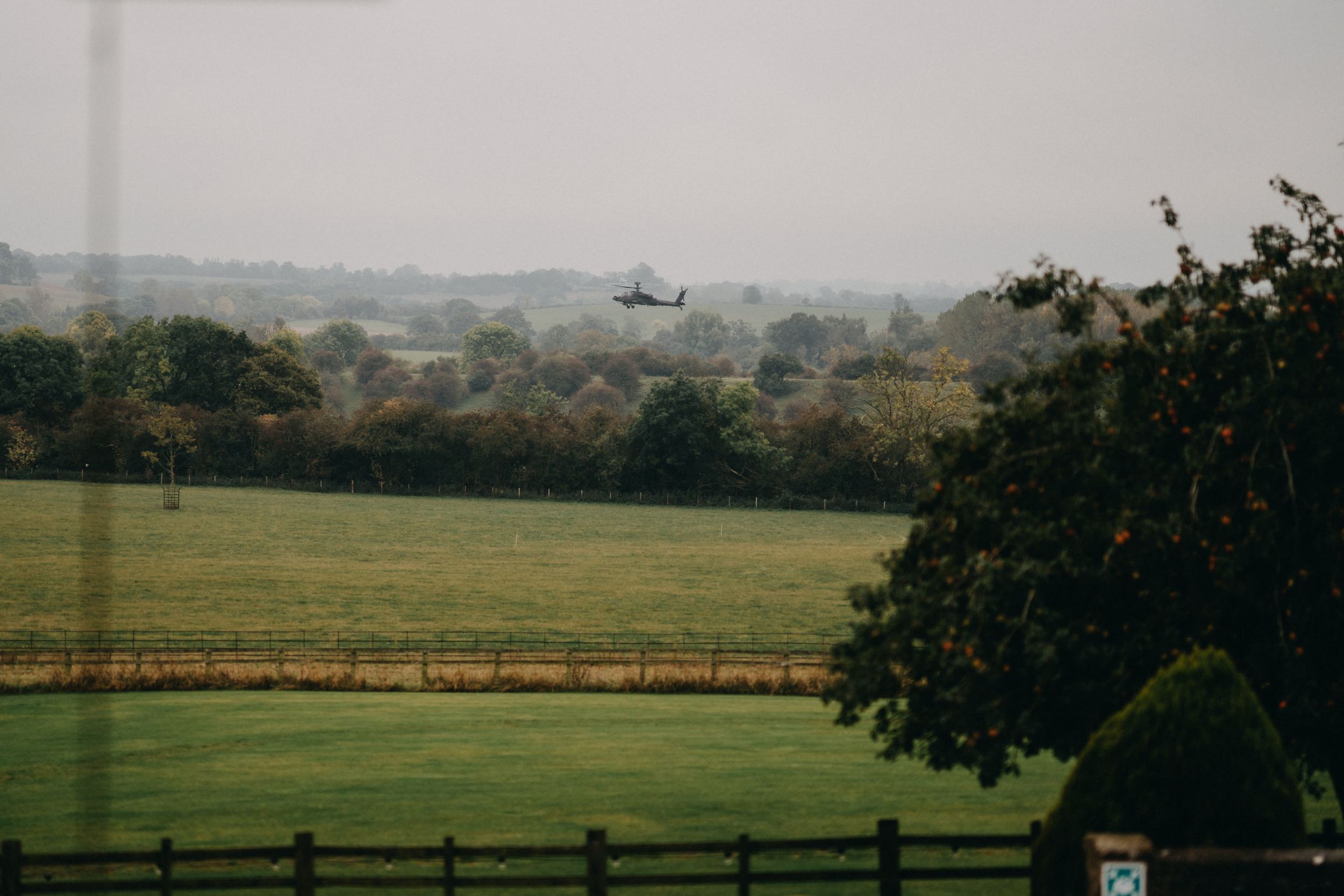 Helicopter flies over scenic countryside landscape with rolling hills and fenced fields.