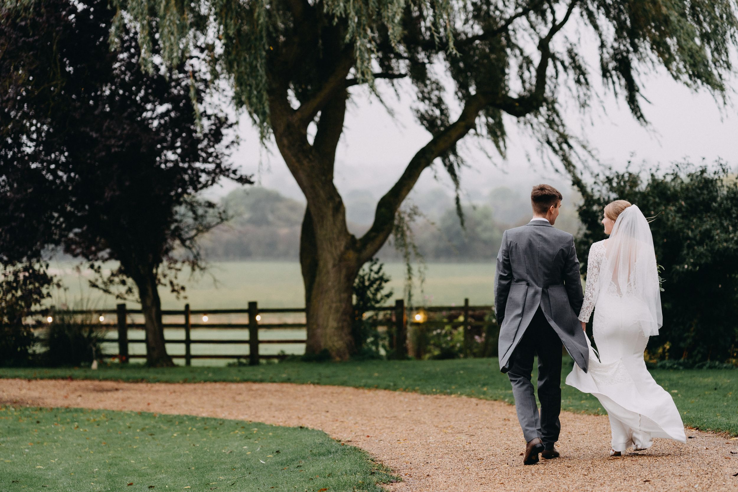Newlyweds walking down a garden path beneath a weeping willow tree.