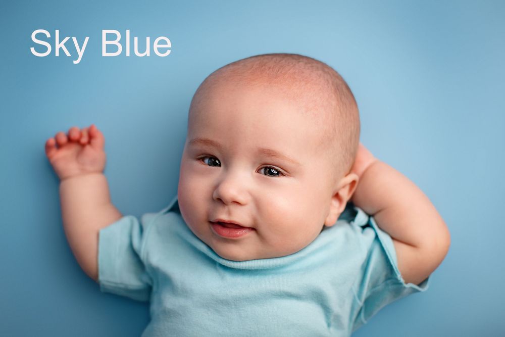 Baby wearing light blue onesie smiles while lying against sky blue background.
