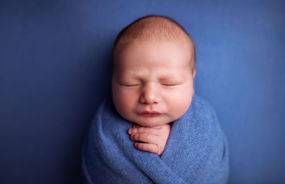 Peaceful newborn wrapped in soft blue blanket sleeping soundly against blue backdrop during professional photo session.