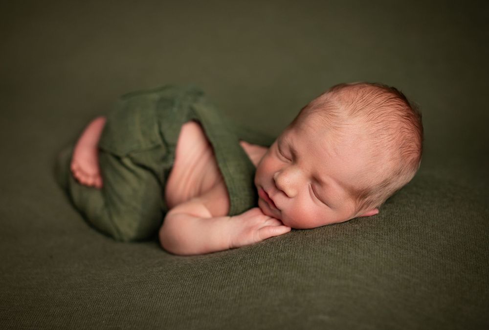 Sleeping newborn baby wearing green wrap curled up on dark green textured background in studio setting.