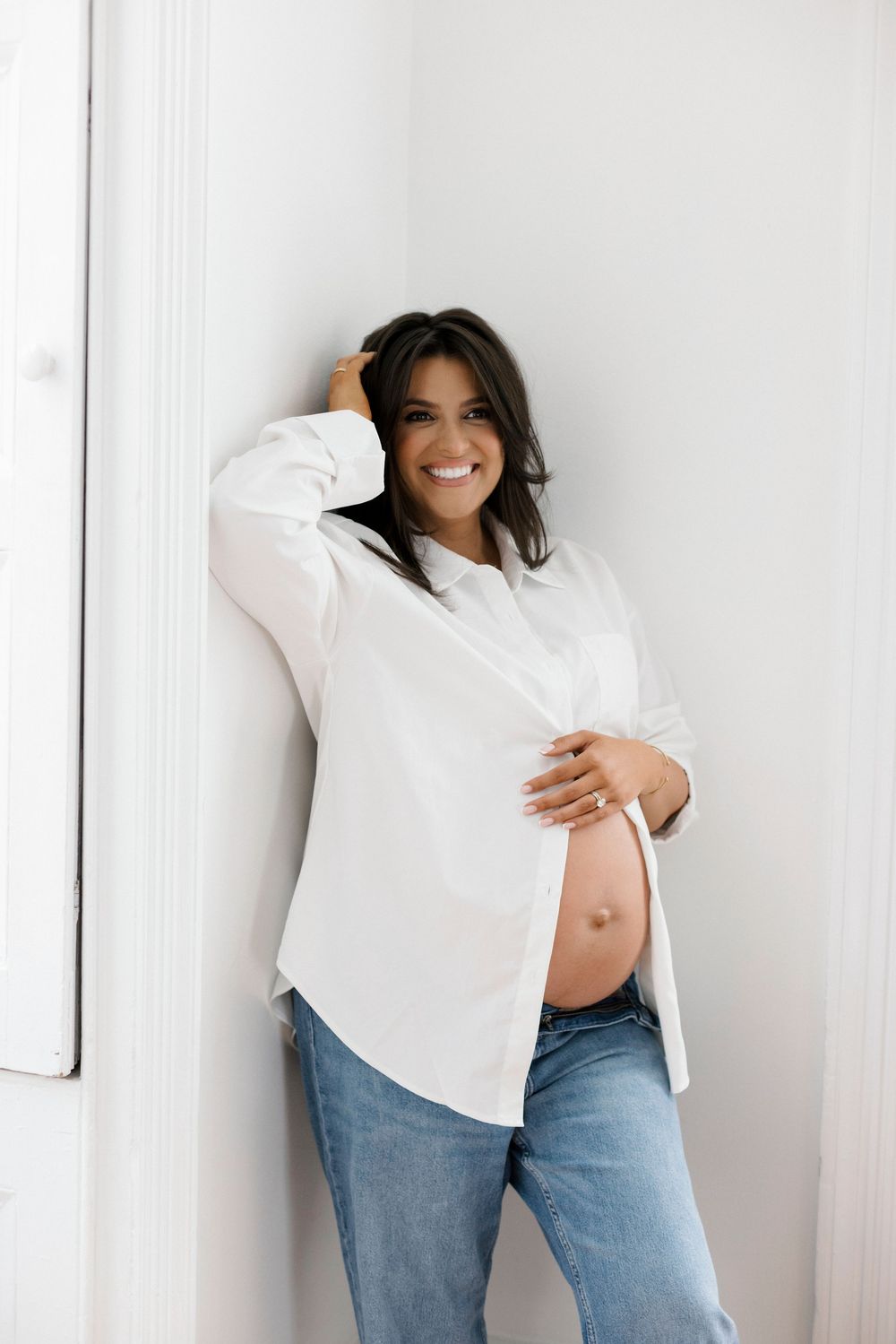 A pregnant woman in a white button-down shirt and jeans poses against a white wall with a joyful expression.