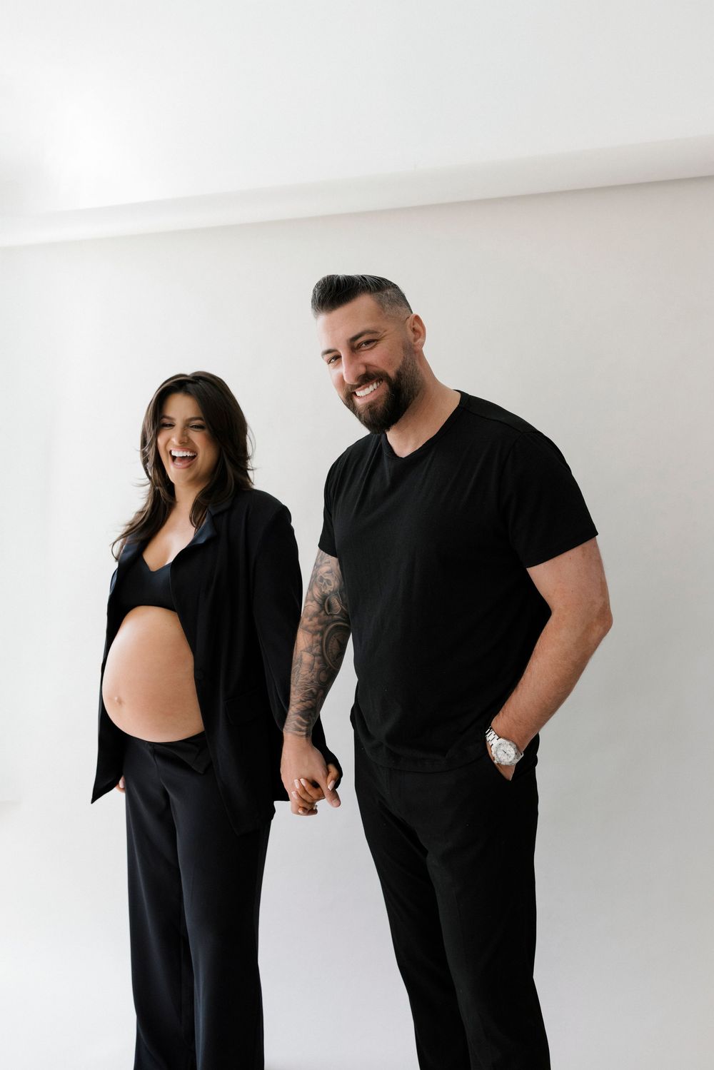 A couple in matching black outfits poses against a white wall during a maternity photoshoot.