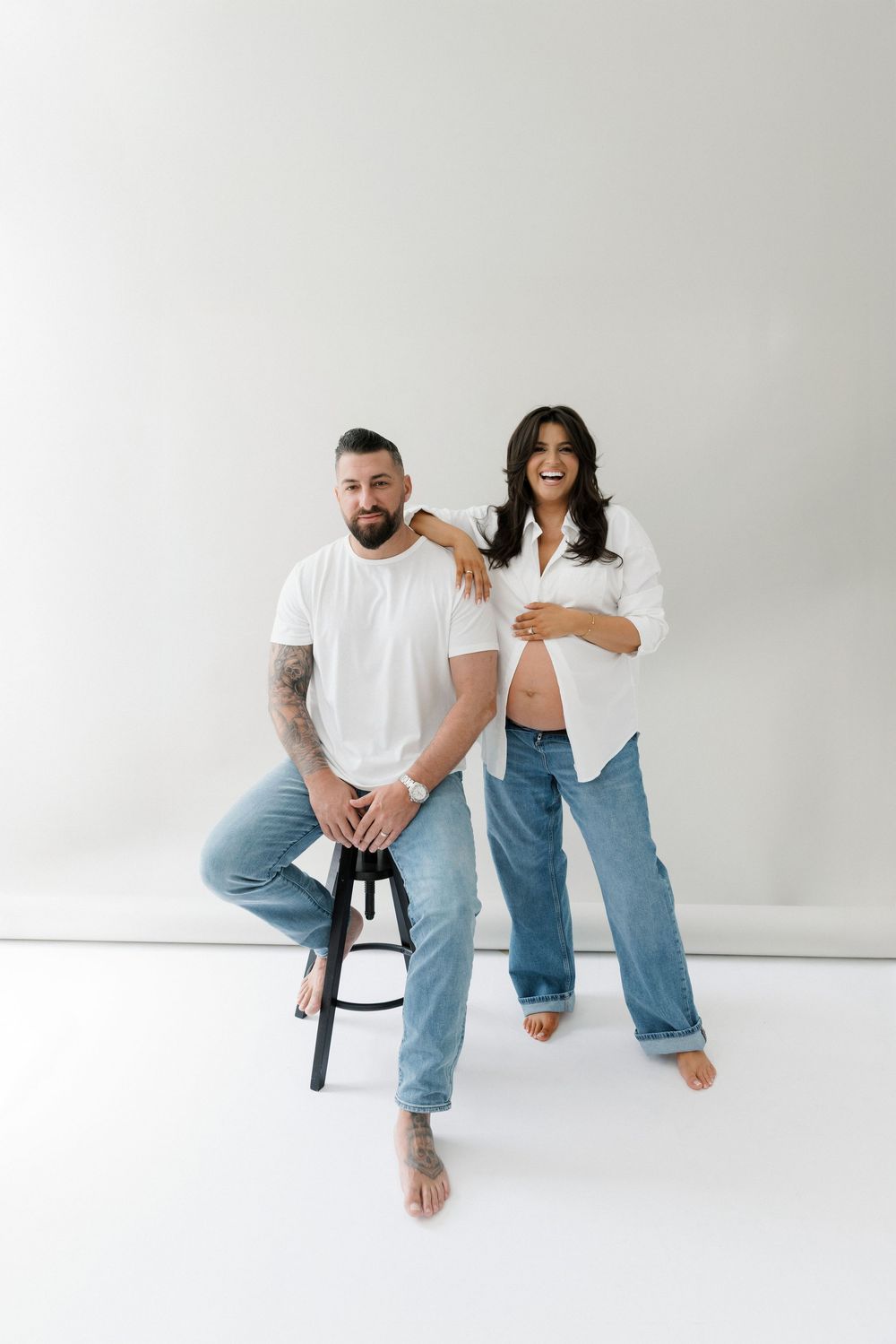 Two people in casual white shirts and jeans pose together against a white wall, one seated on a stool.