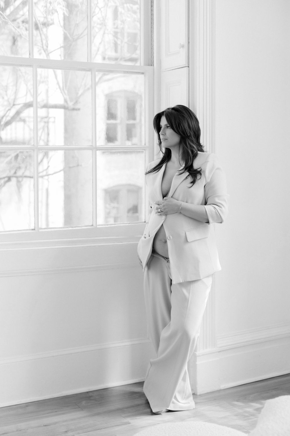 A person in a light business suit gazes pensively through a bright window in a minimalist white interior.