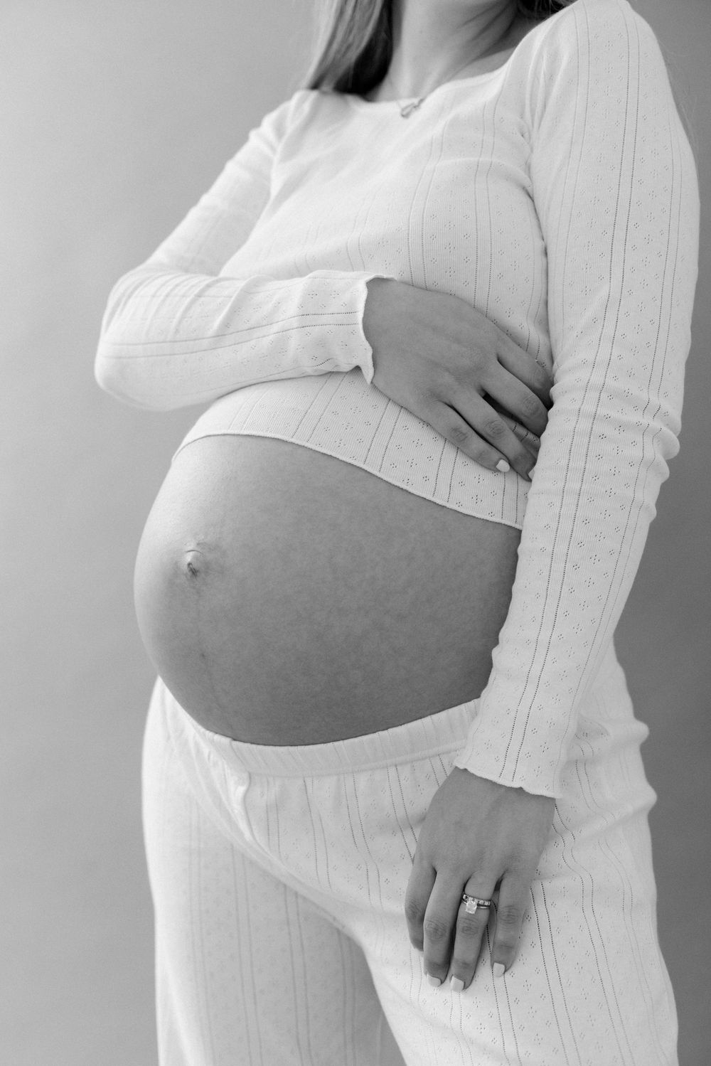 Black and white photo showing pregnant belly in white ribbed loungewear with woman's hand resting on stomach.