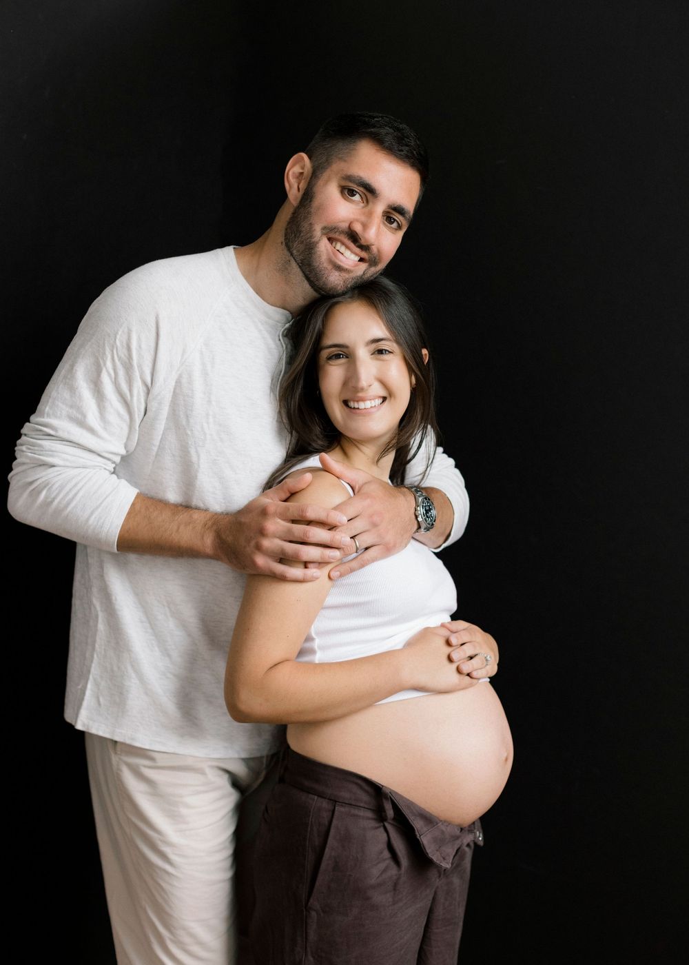 A couple wearing white tops embrace in a maternity photo against a dark background.