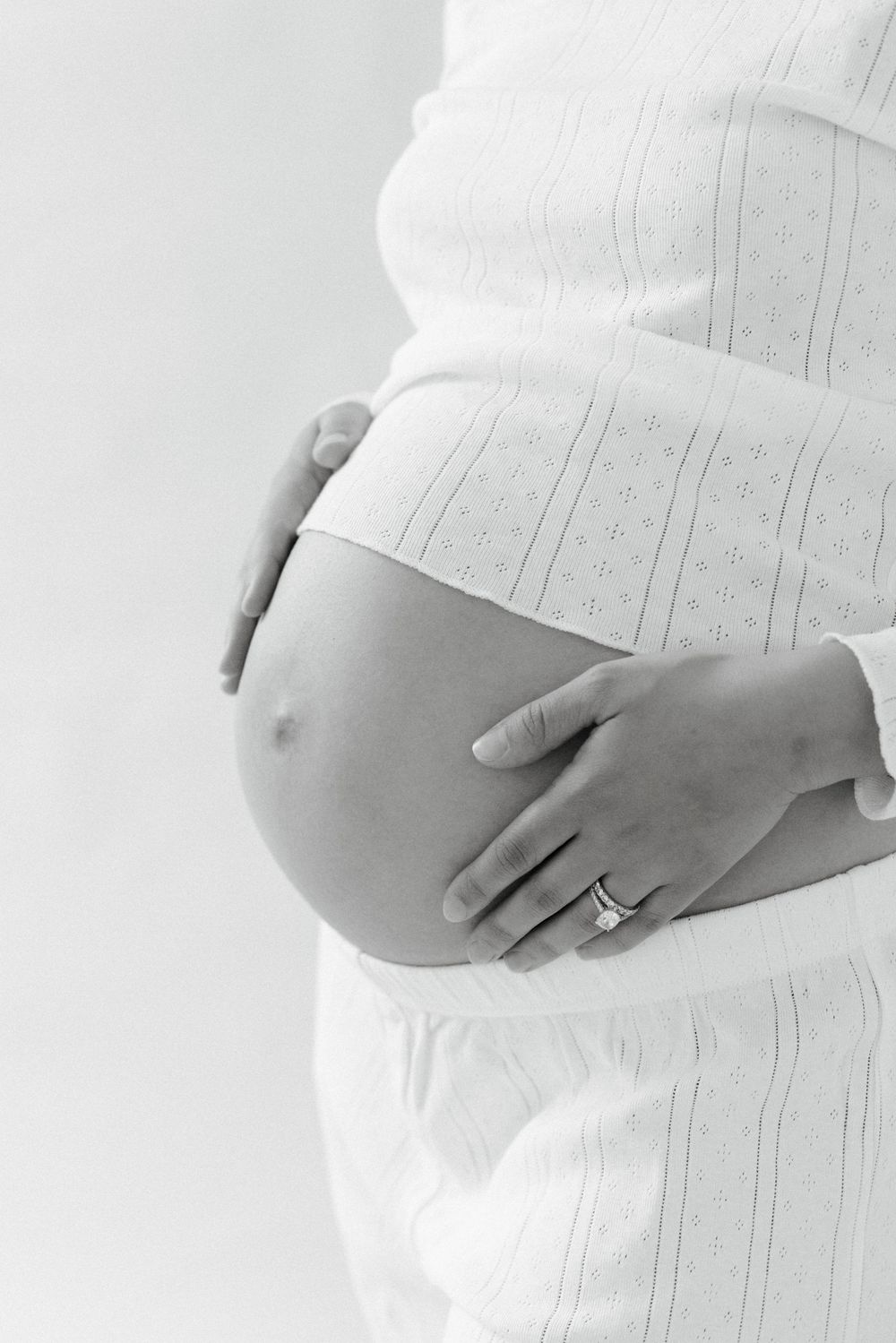 Close-up black and white photo showing hands cradling a pregnant belly wearing a white textured dress with a ring.