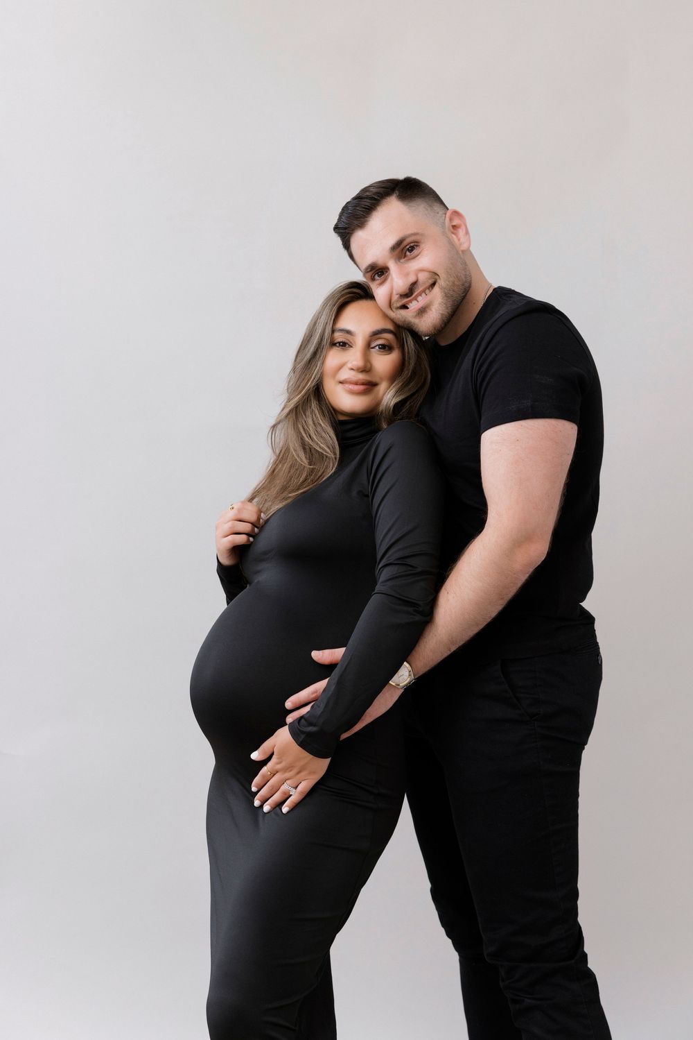 A couple dressed in black embraces during a minimalist maternity photo session against a light background.