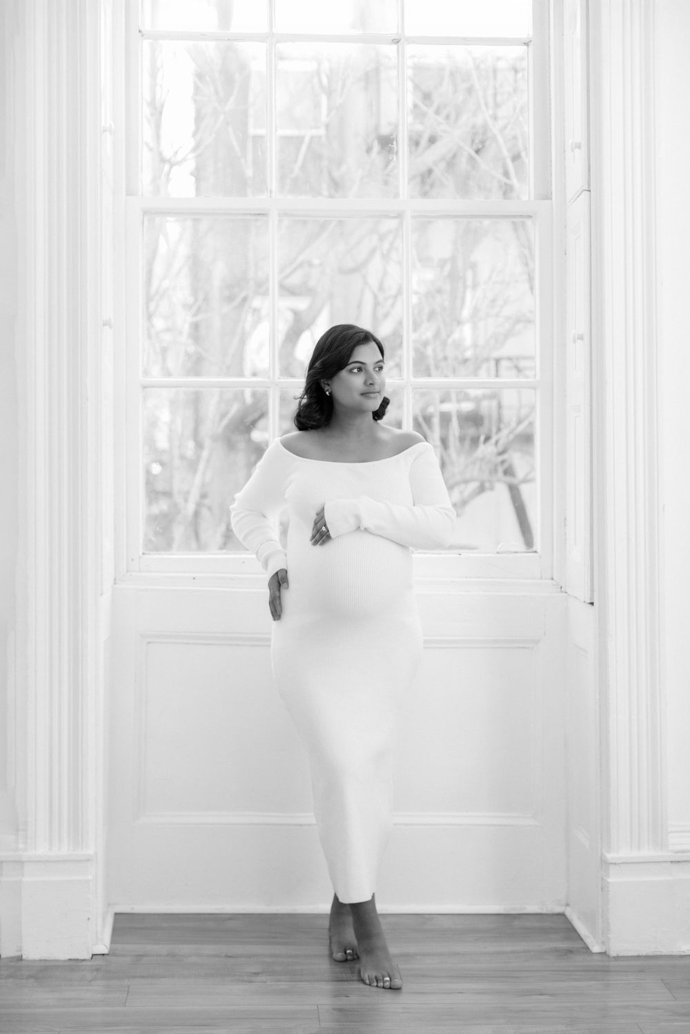 A black and white portrait of someone wearing an elegant off-shoulder white dress posing by a bright window.