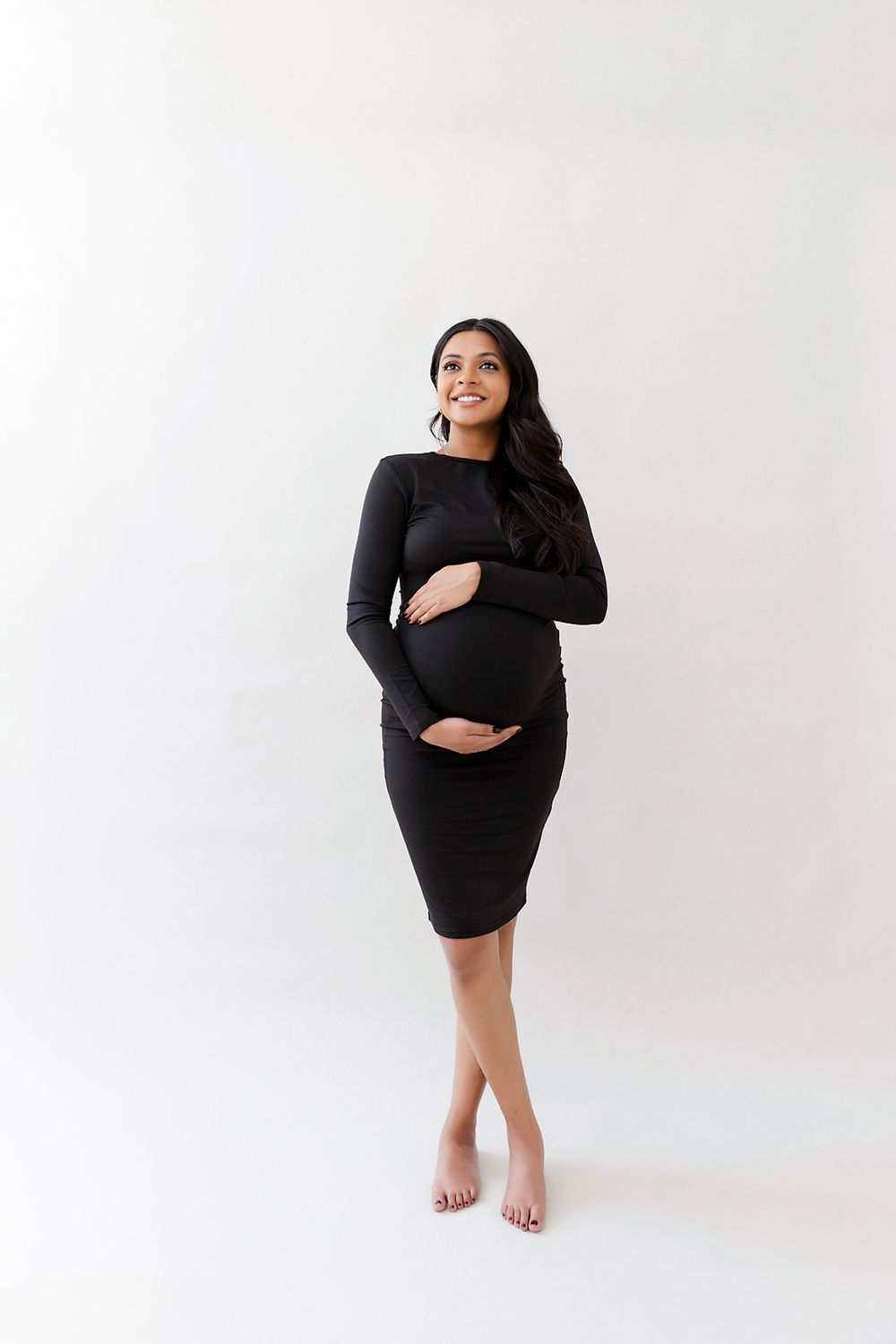 Expectant woman in black long sleeve dress poses barefoot against white backdrop for maternity photoshoot.