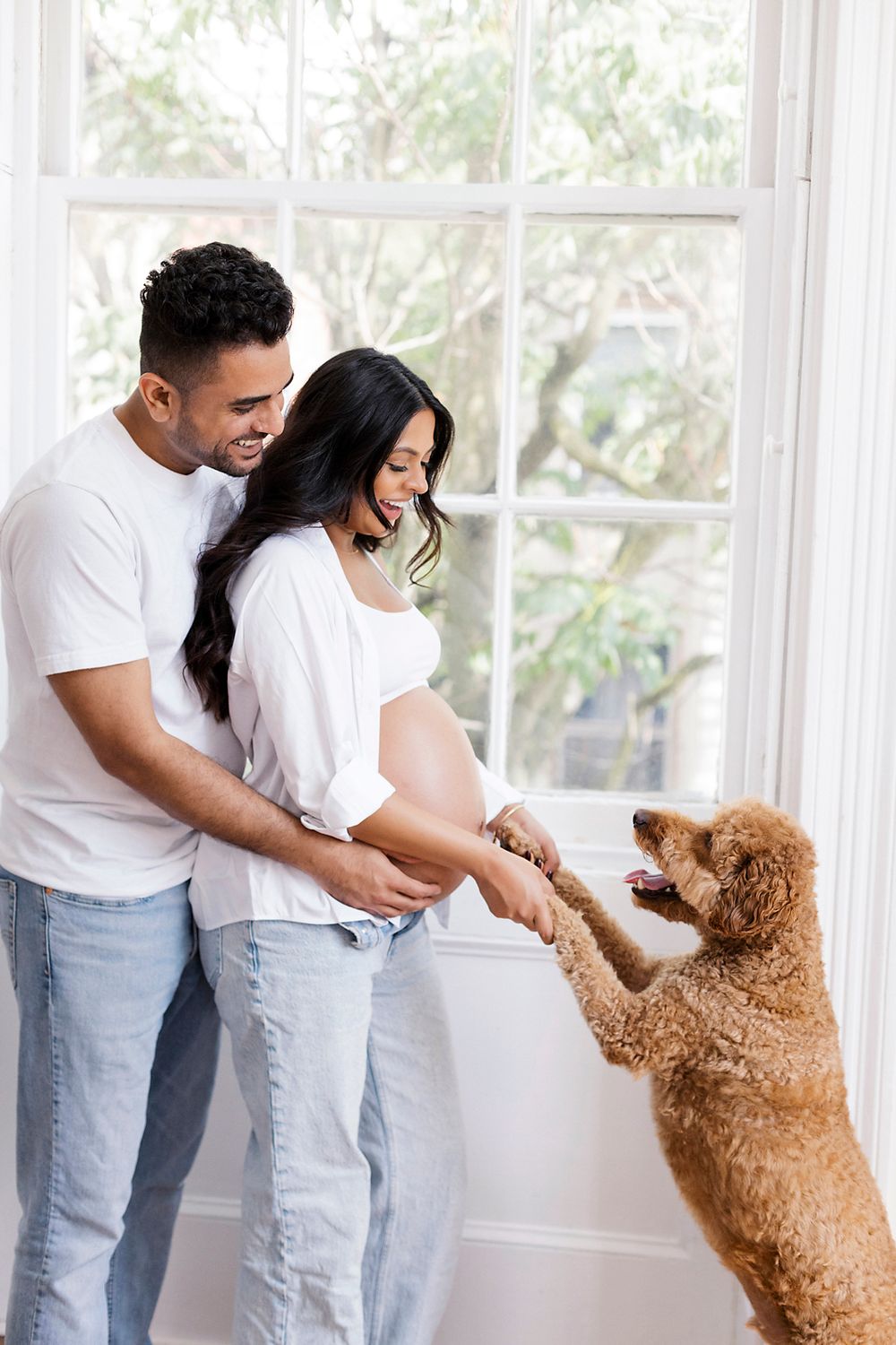 A couple with their goldendoodle dog looking out a sunlit window in their home together.