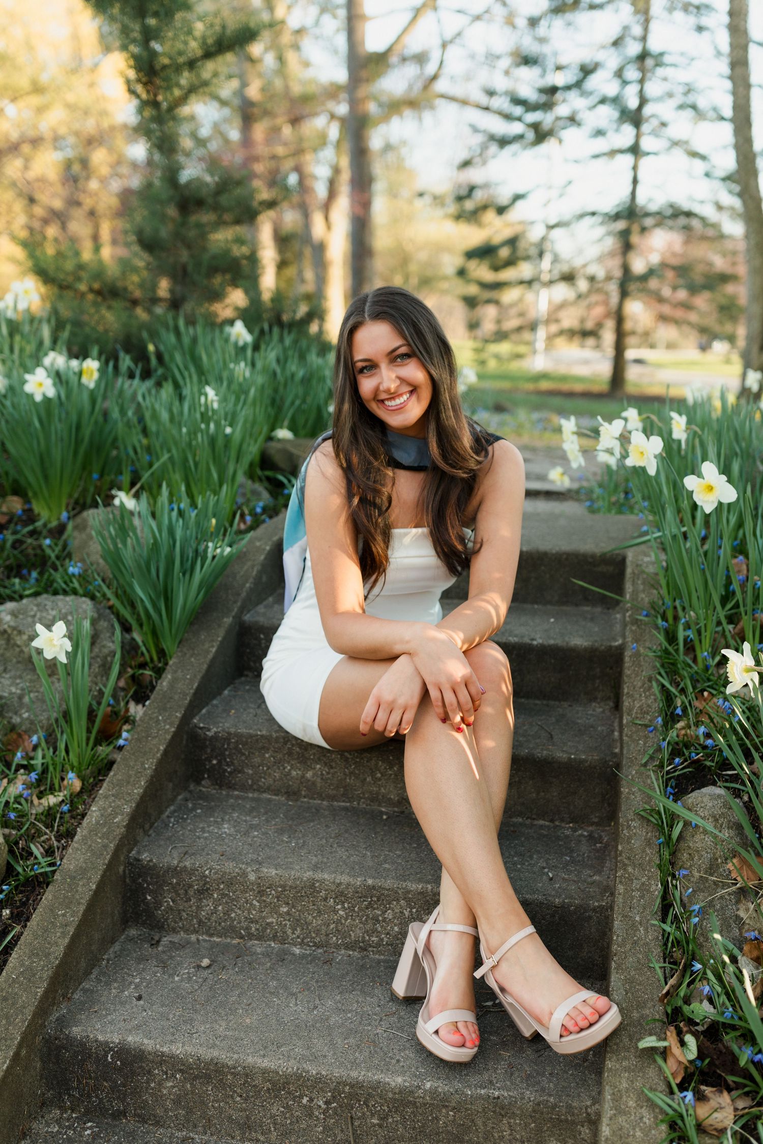 Graduate seated on stone steps surrounded by blooming white daffodils in spring garden setting.