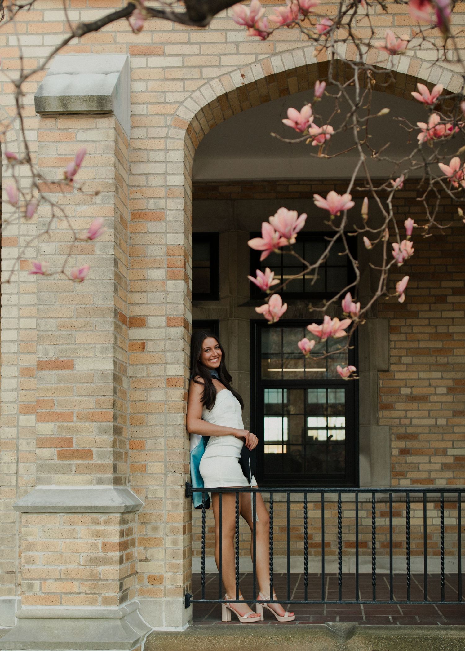 Graduate poses in archway framed by blooming pink magnolia flowers against brick building.