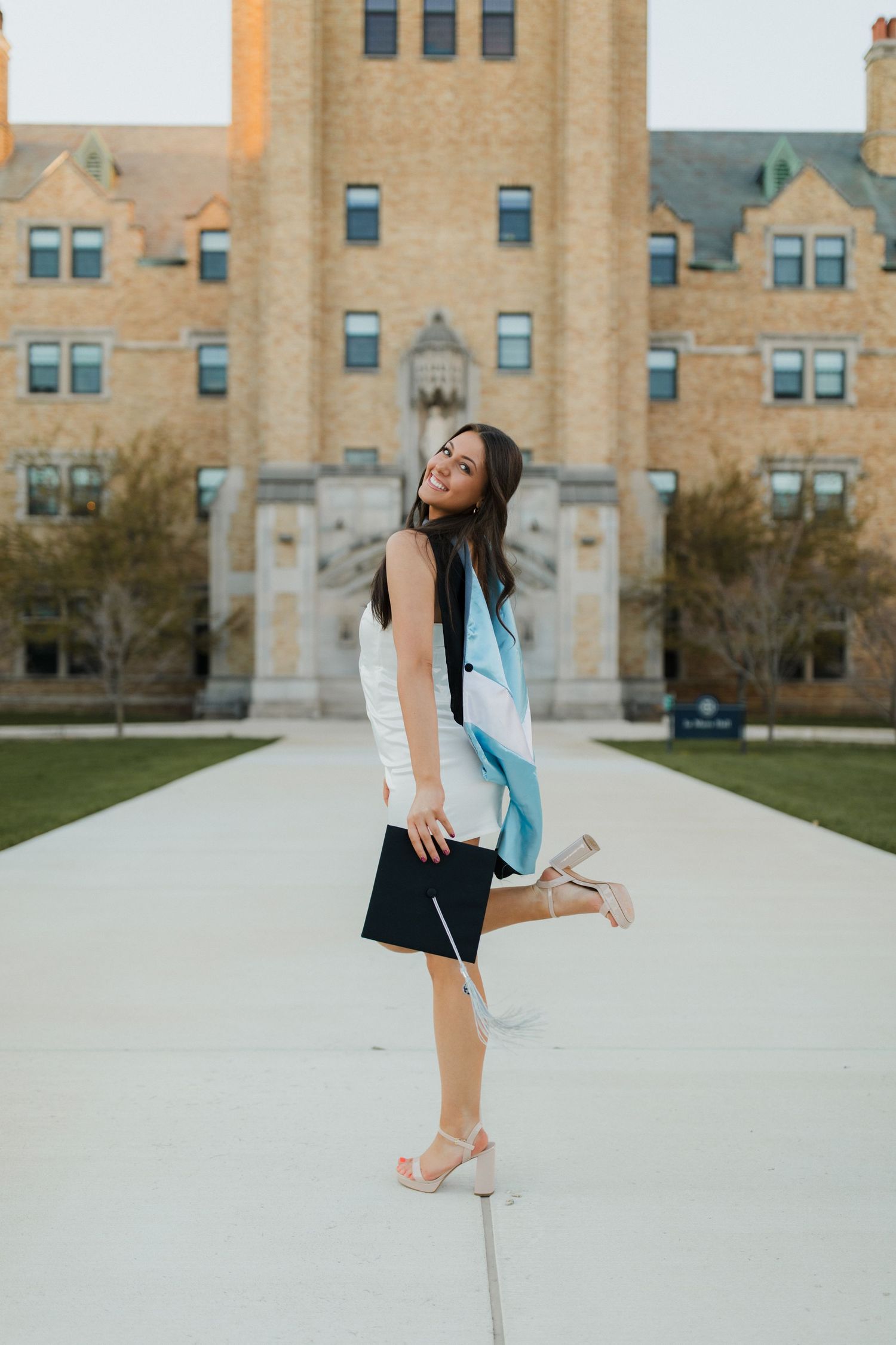Graduate celebrating on campus sidewalk with historic university building in background.