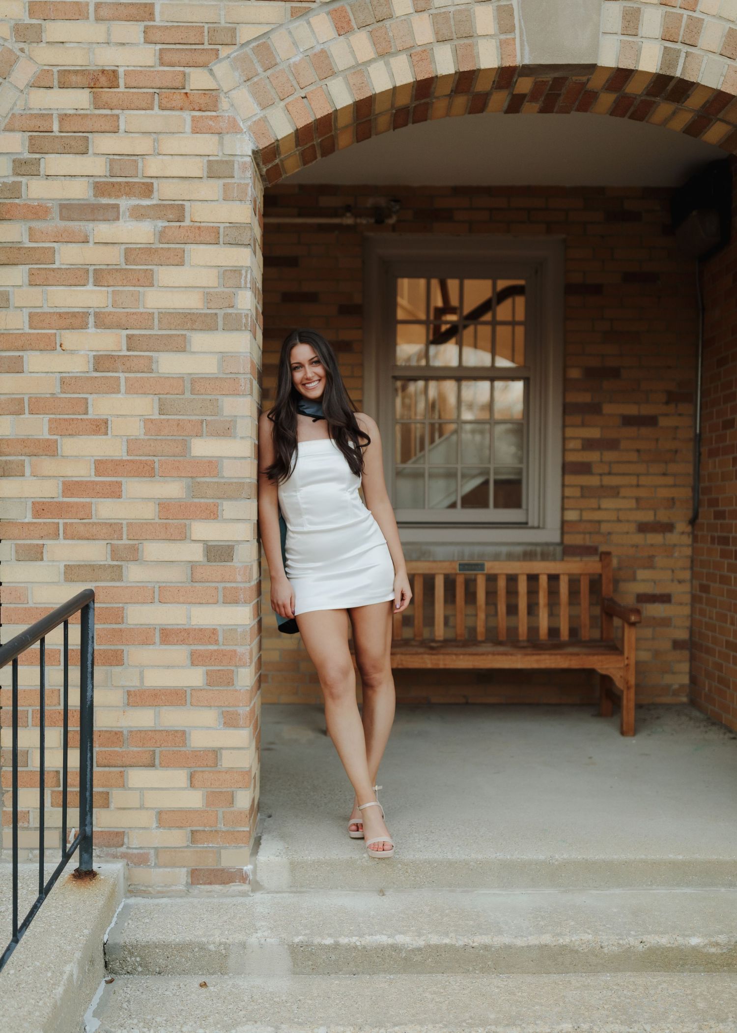 Elegant white mini dress and high heels photographed in front of a brick archway with wooden bench.