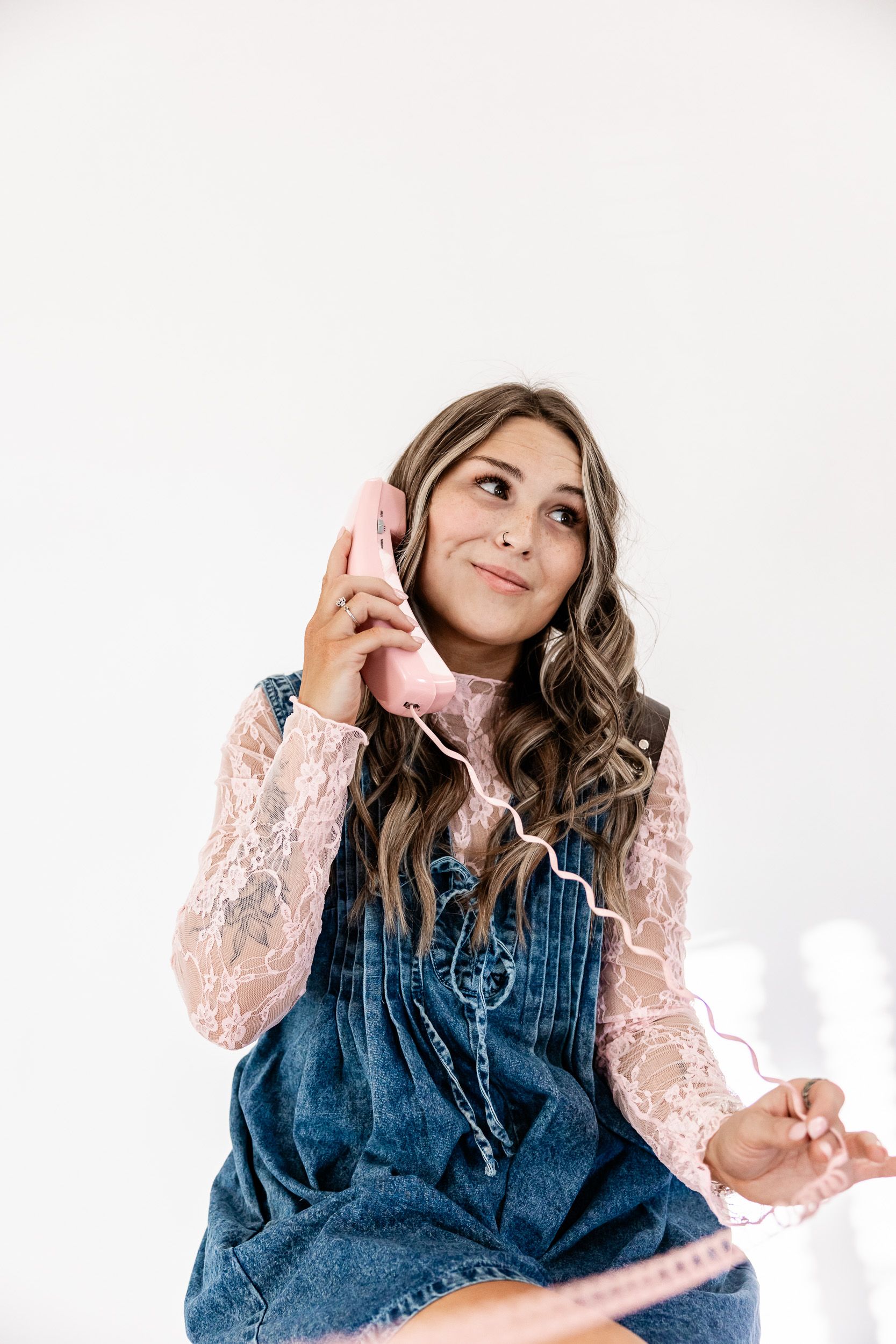 Wyoming Photographer
Person wearing denim overalls and lace blouse talking on pink retro telephone with spiral cord against white background