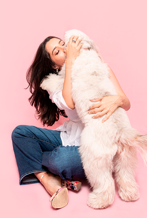 Person in denim hugging fluffy white dog against pink background.