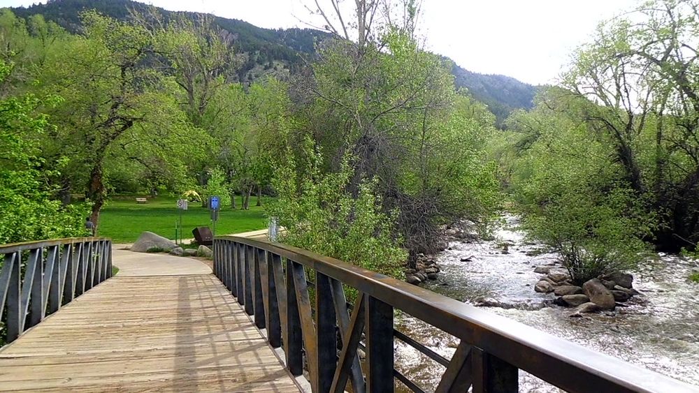 A wooden footbridge crosses over a flowing stream surrounded by lush green trees and mountains in the background.