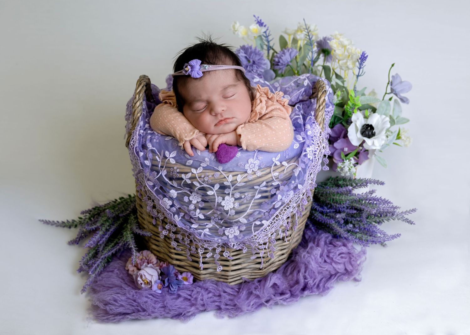 Newborn photography setup with purple flowers and woven basket on soft lavender fabric.