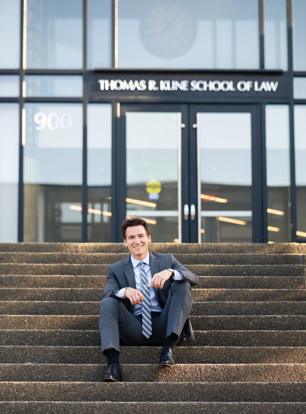 A professional wearing a dark suit and tie sits casually on outdoor concrete steps outside a law school building.