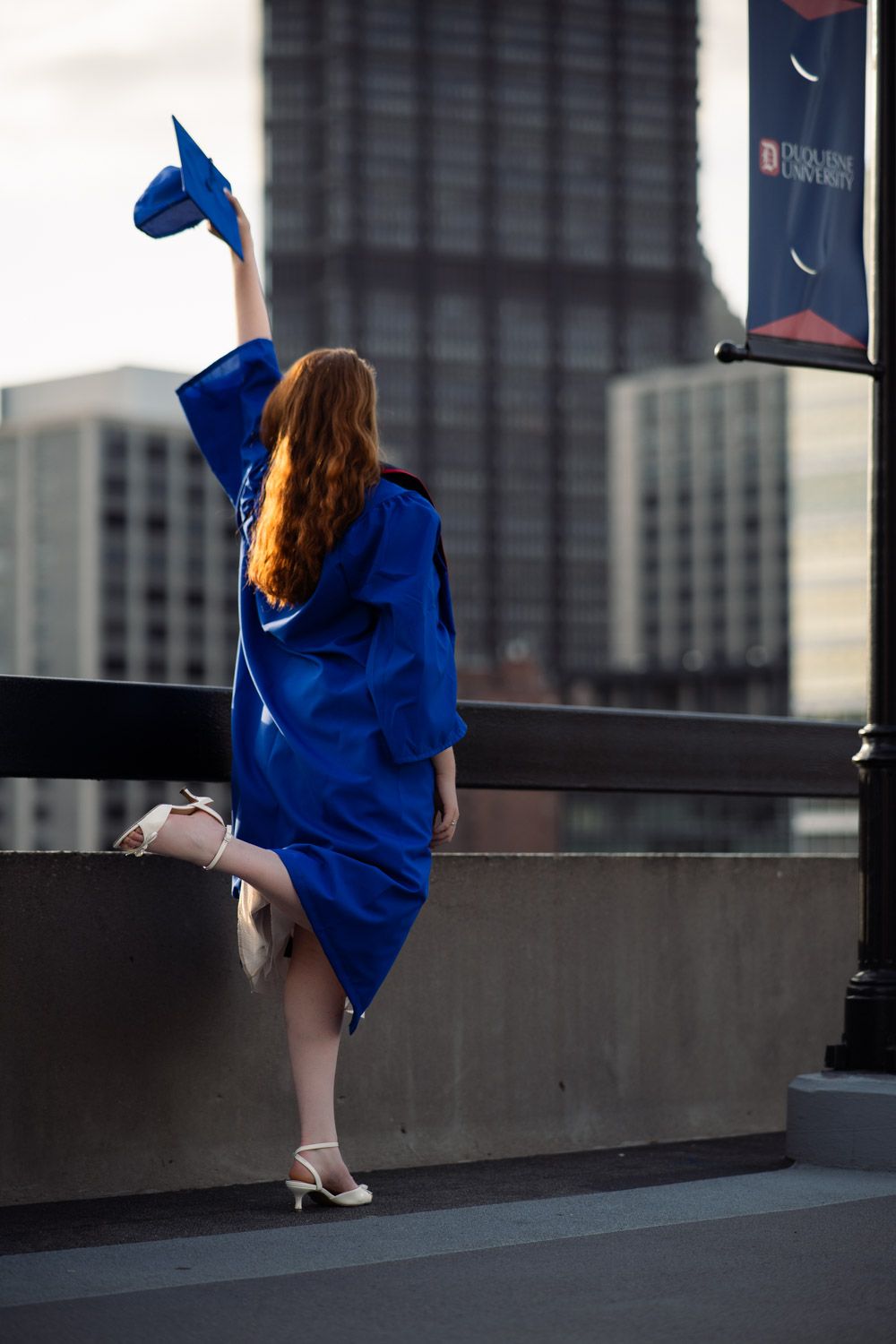 Graduate in bright blue cap and gown celebrates on rooftop with city buildings in background during graduation photoshoot.