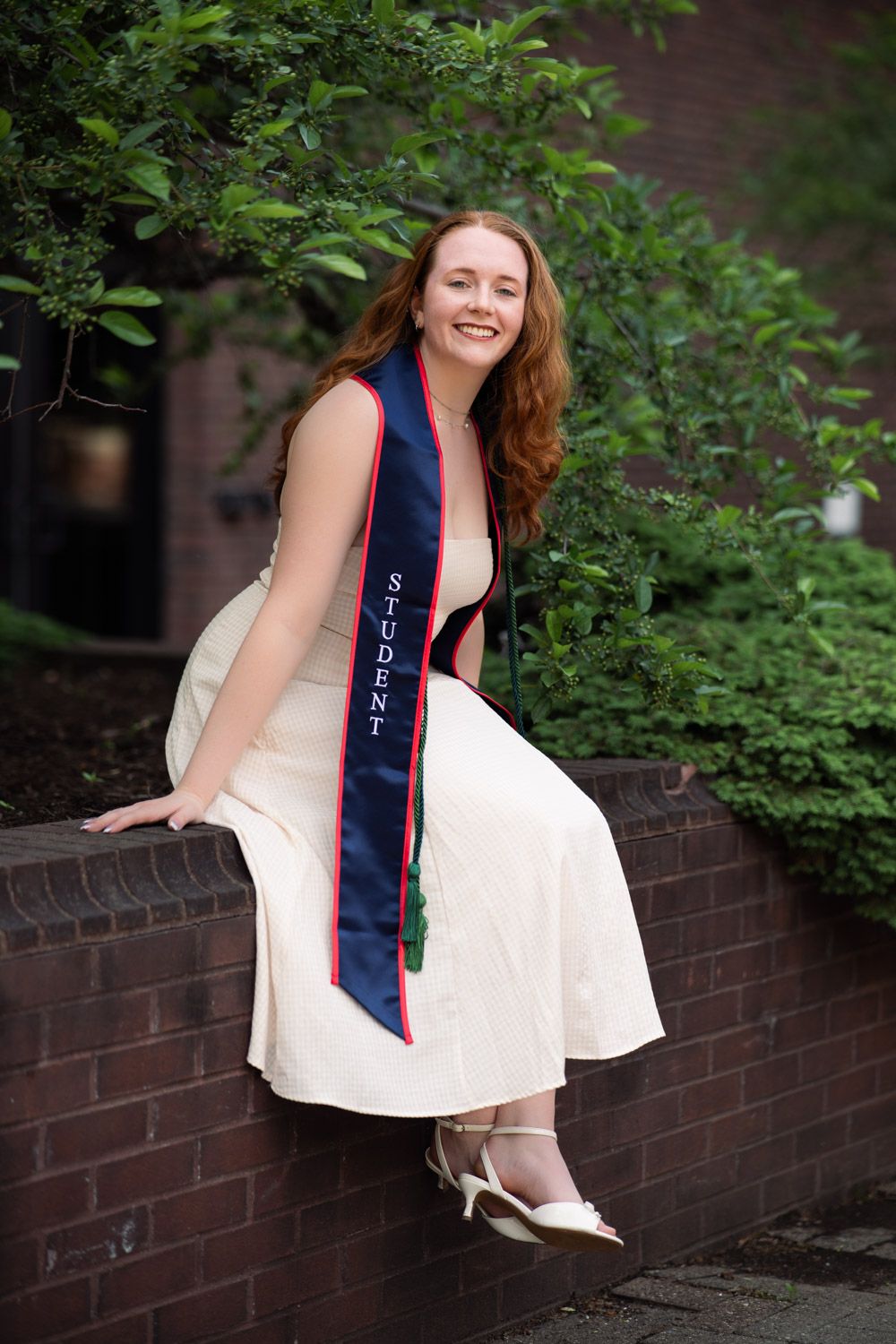 Graduate wearing a white dress and blue sash sits on brick wall surrounded by greenery in outdoor setting.