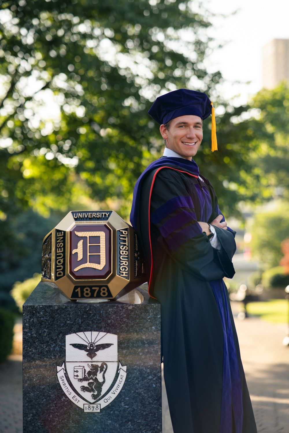 Graduate in academic regalia poses by stone monument with university logo and crest on a sunny day.