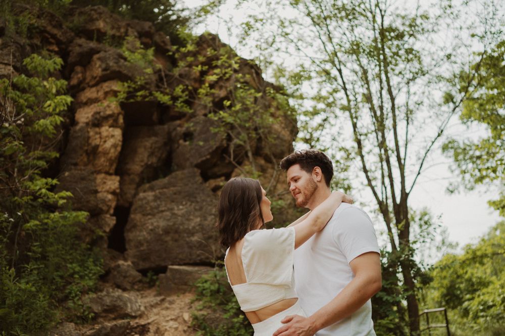 Two people share a tender moment against a natural rock formation backdrop in a wooded area.