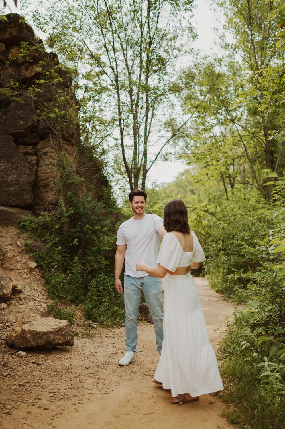 A couple walks hand in hand along a dirt trail surrounded by lush greenery and rock formations.