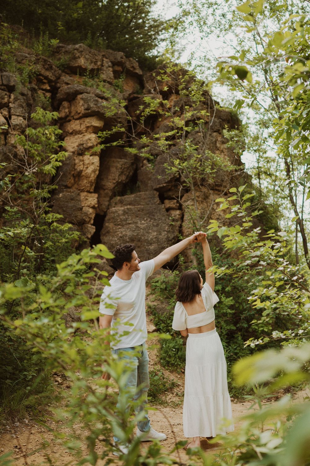 Two people dance playfully on a dirt path with dramatic rock walls and green trees surrounding them.