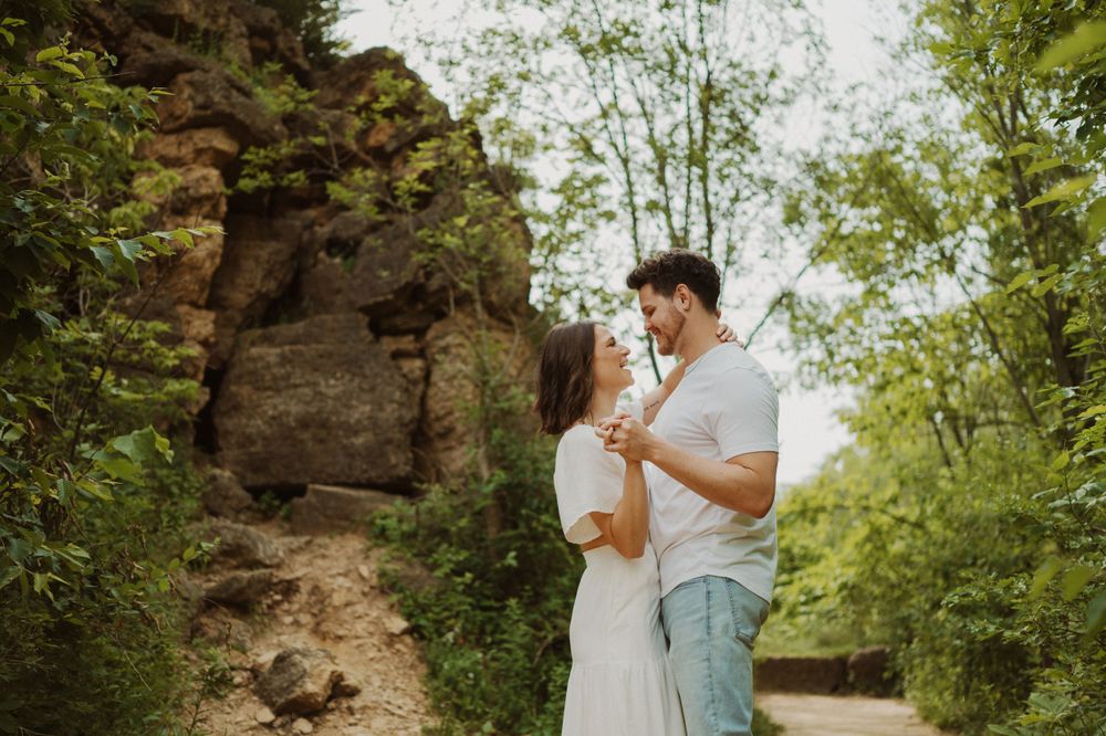 Two people share a romantic embrace on a sunlit trail with rocky cliffs in the background.