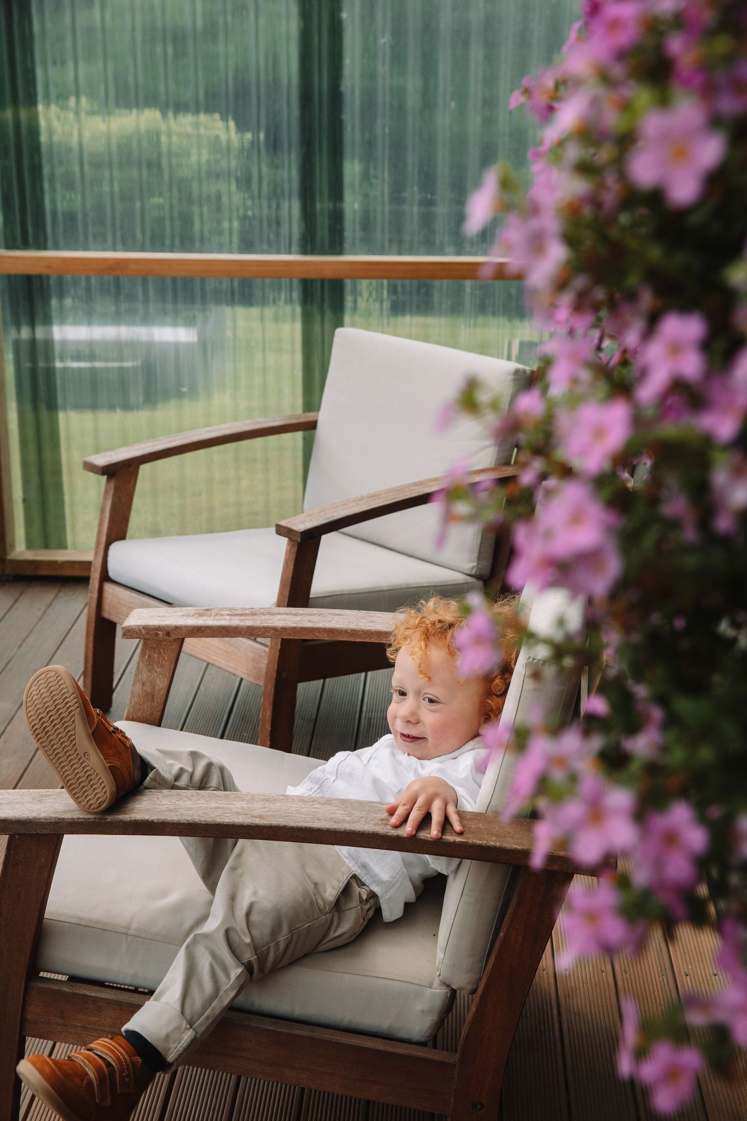 Child relaxing in a modern wood chair next to blooming pink flowers on a deck.