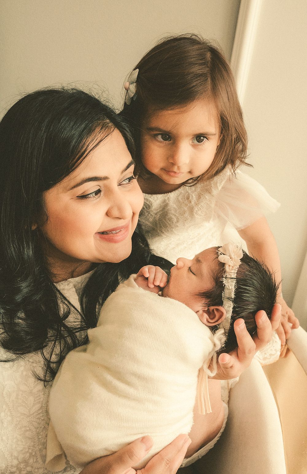 Two figures in white clothing share a tender moment with a newborn baby wrapped in a cream-colored blanket.